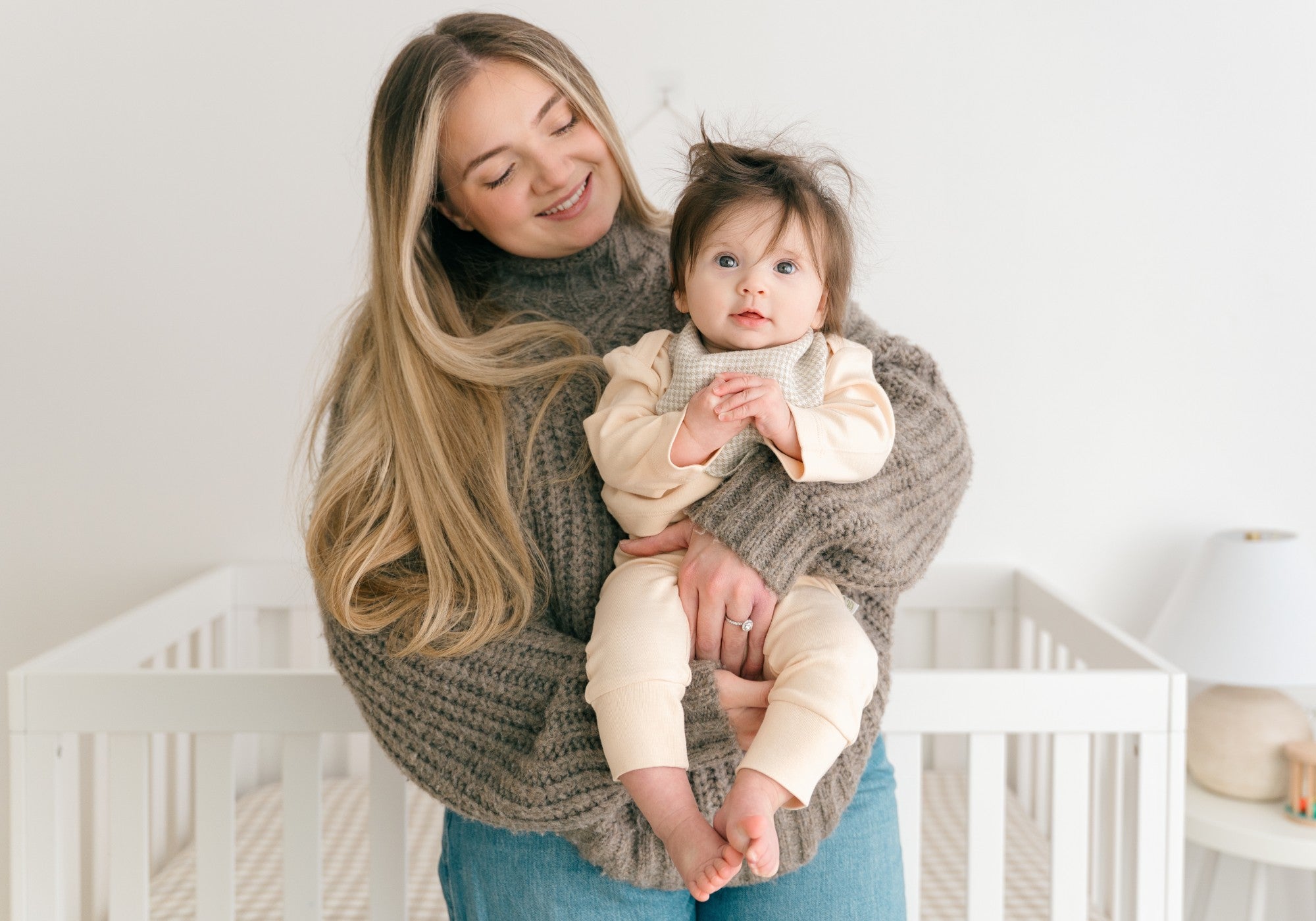 A smiling mom holding a baby wearing wearing long sleeve shirt and fold-over pants in rosy peach