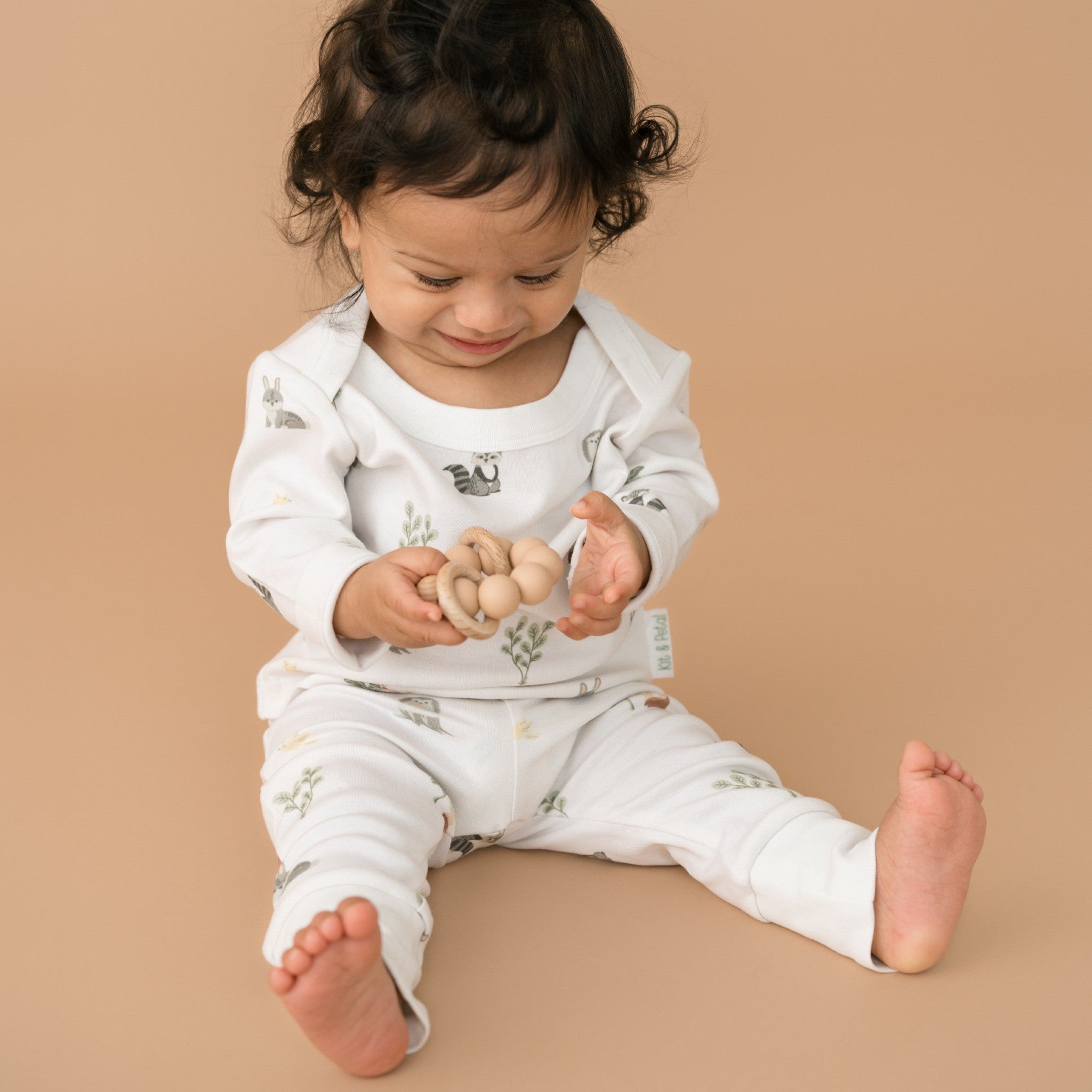 Toddler boy holding a wooden toy while wearing a Kit & Petal long sleeve shirt and fold-over pants in Forest Friends animal print.