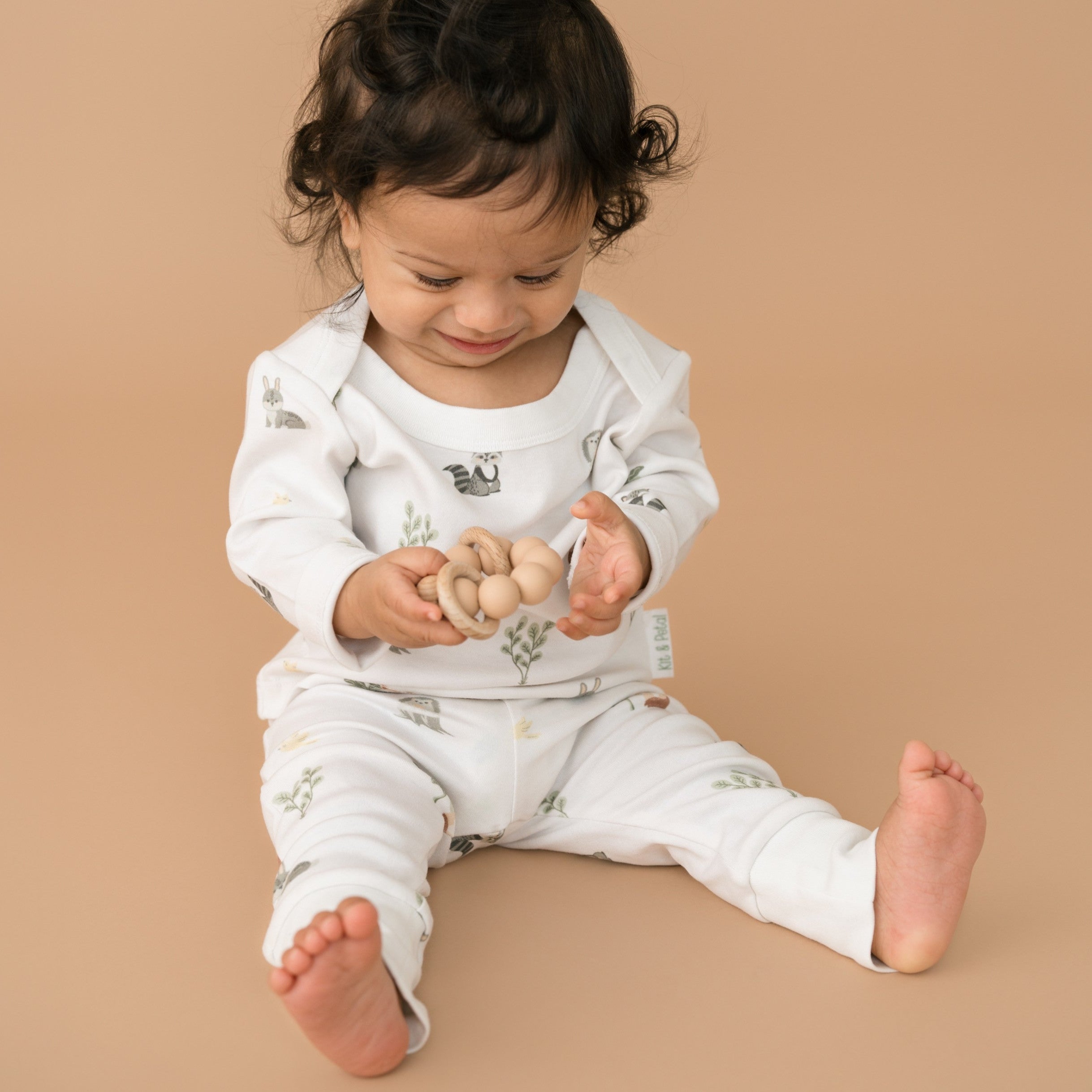 Toddler boy holding a wooden toy while wearing a Kit & Petal long sleeve shirt and fold-over pants in Forest Friends animal print.