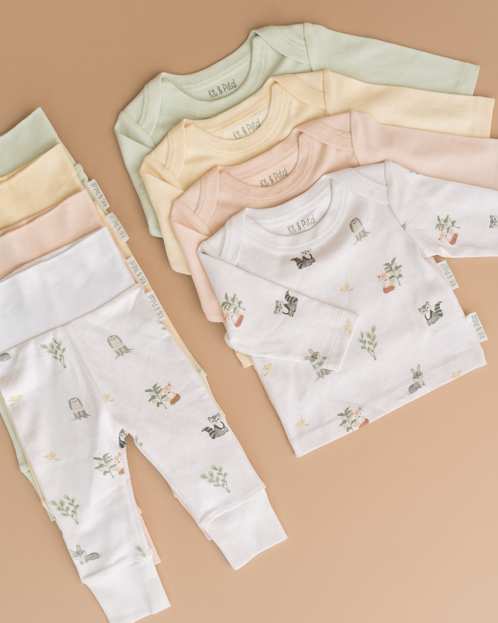 A stack of four long sleeve shirts in four different colorways, and a stack of four fold-over pants in four different colorways. Forest friends animal print, rosy peach, sunny yellow and leafy green.