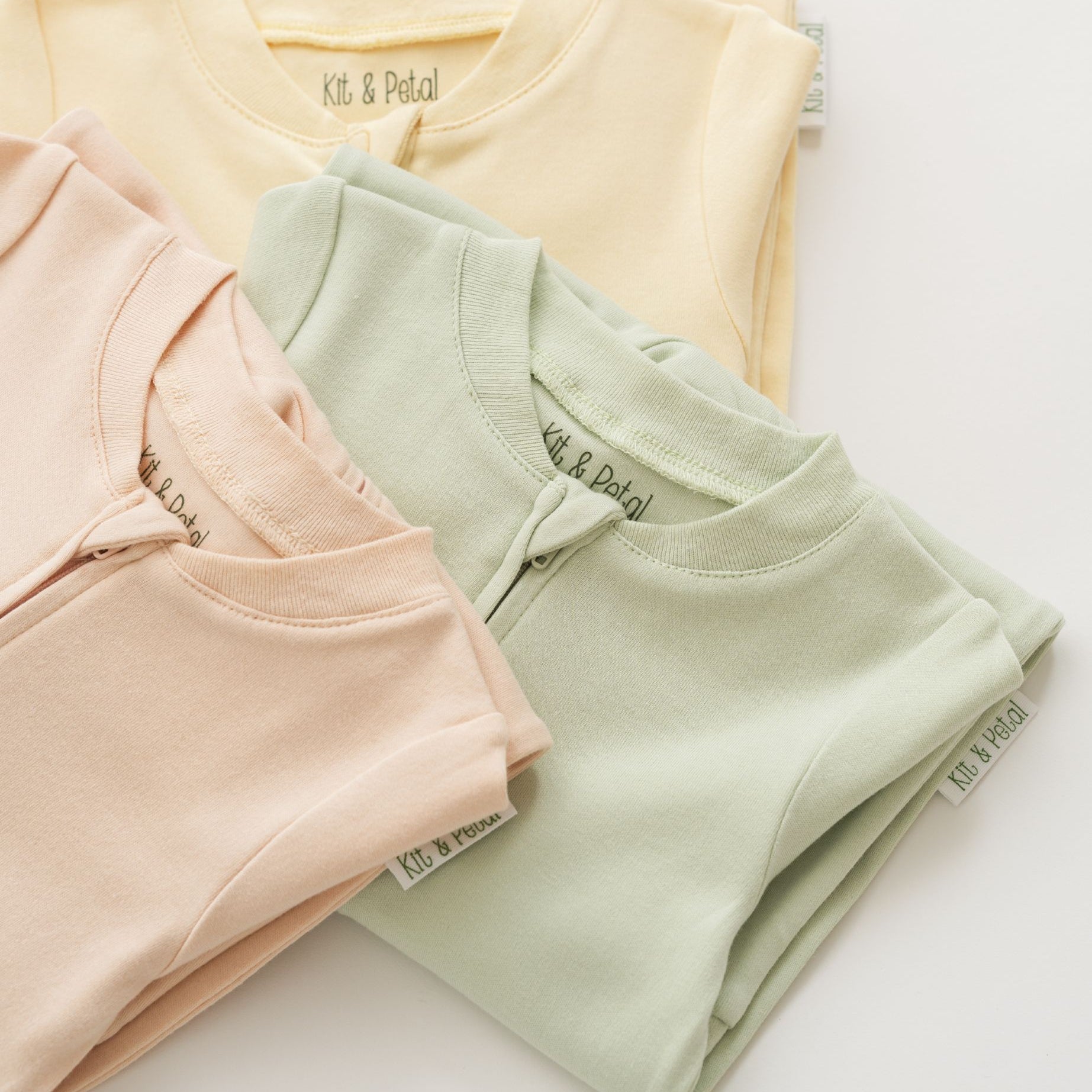 Stack of organic cotton baby long sleeve rompers in leafy green, rosy peach, and sunny yellow, with leafy green in focus