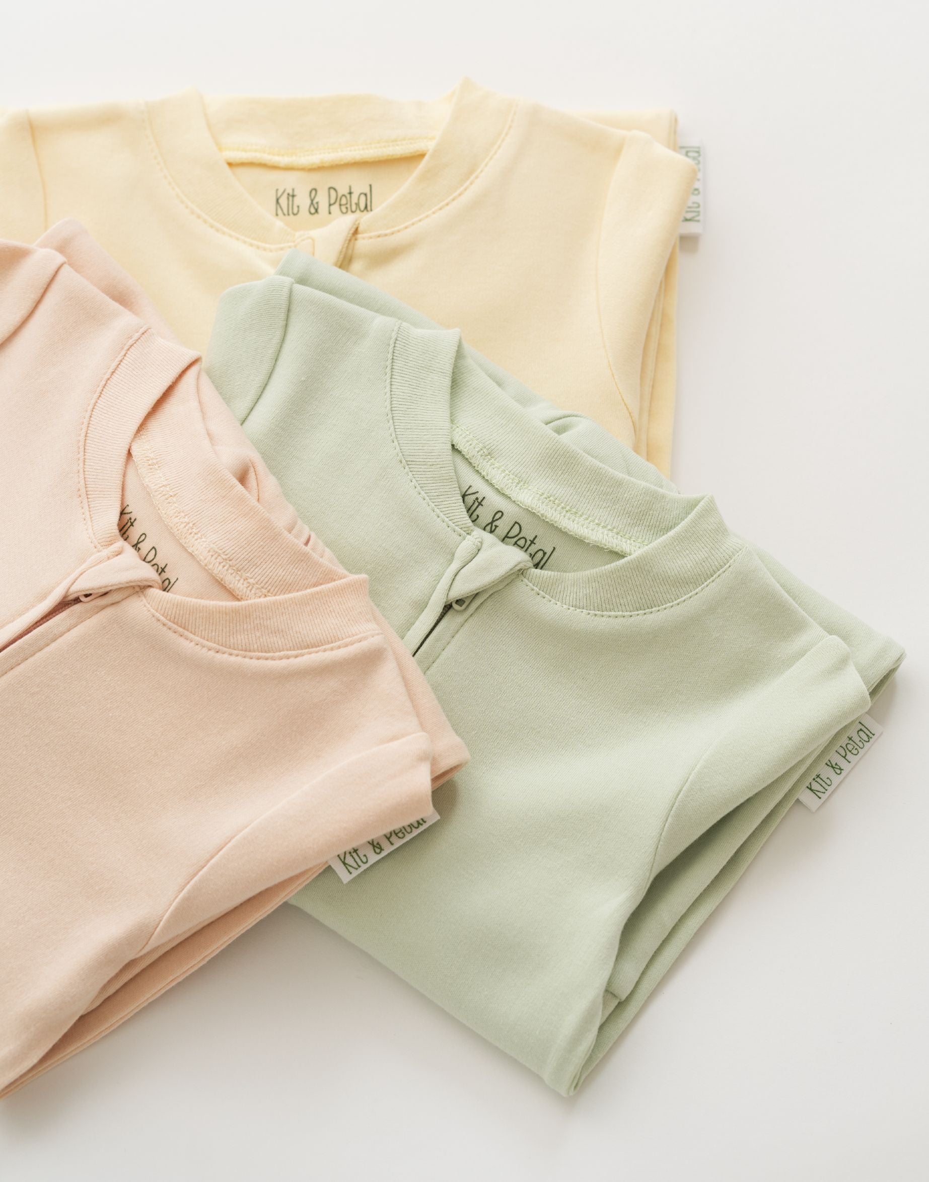 Stack of organic cotton baby long sleeve rompers in leafy green, rosy peach, and sunny yellow, with leafy green in focus