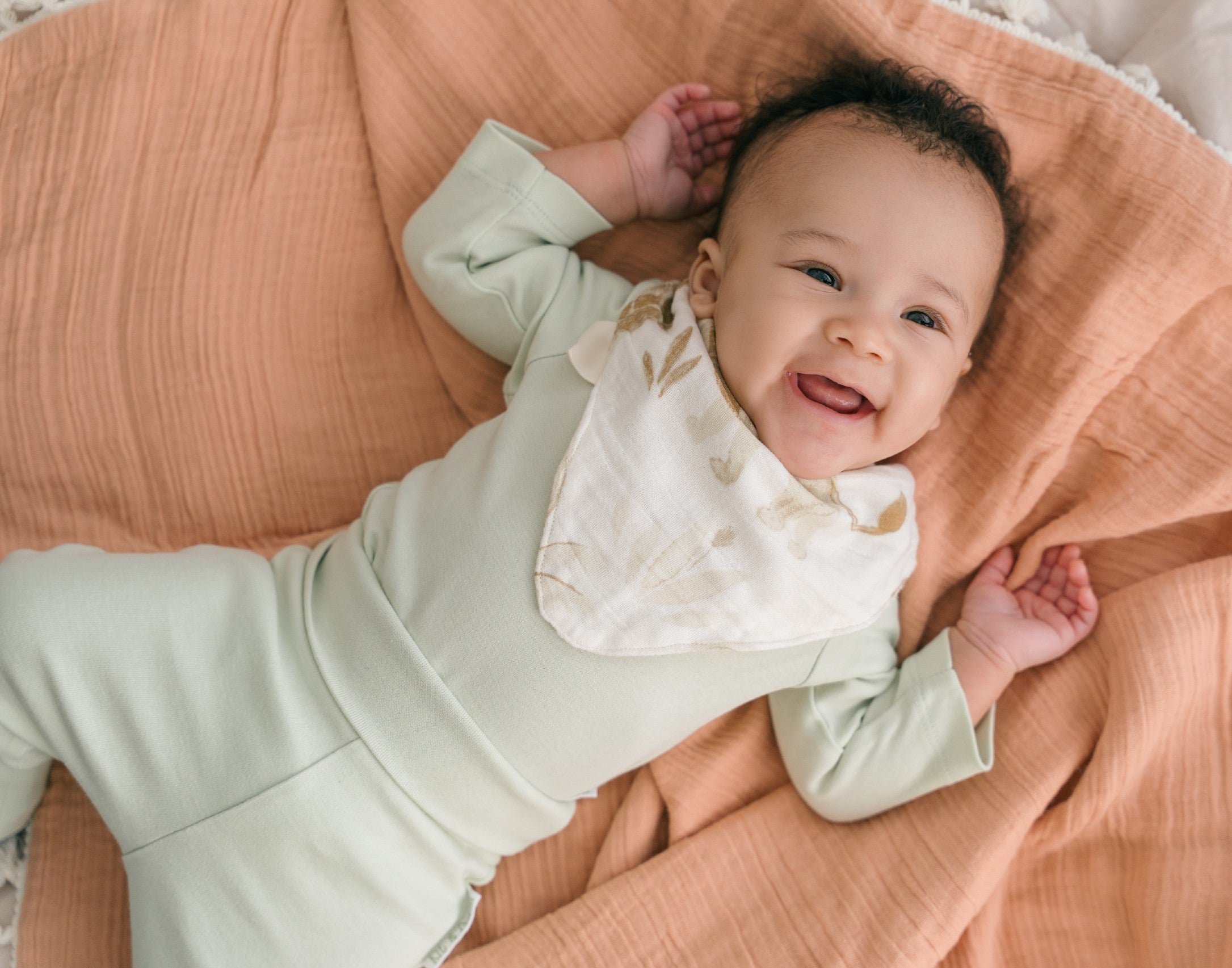 Smiling baby boy wearing a Leafy Green organic cotton long sleeve bodysuit and fold-over pants, laying on a pillow.