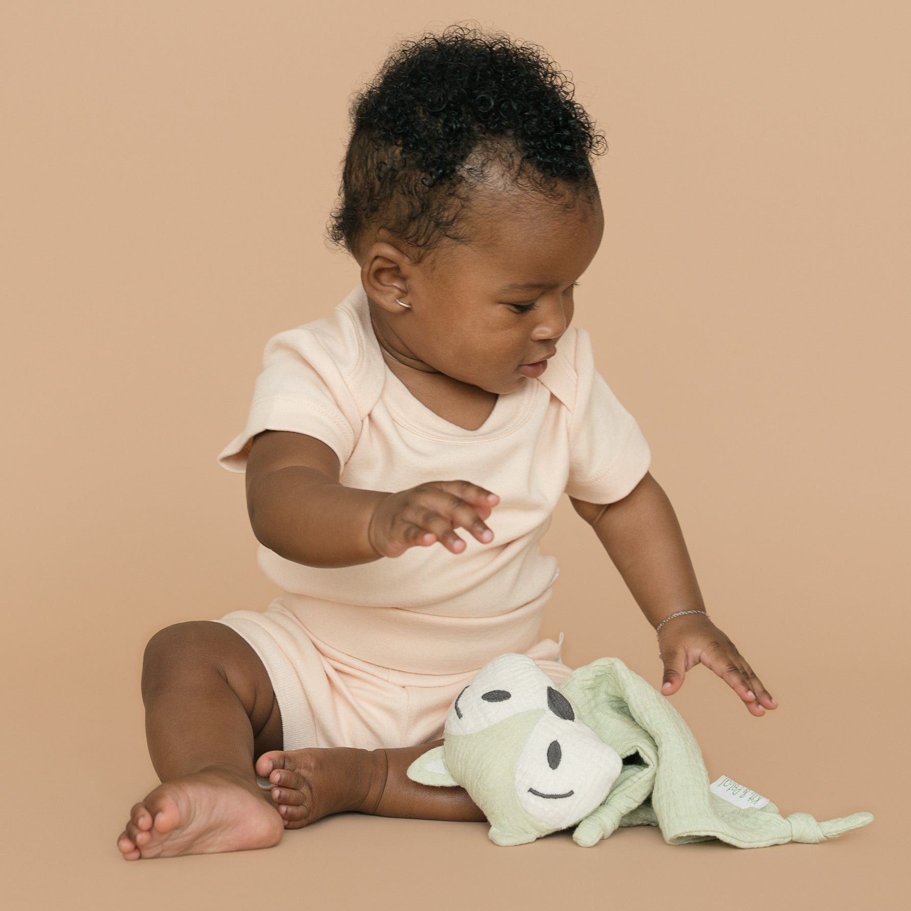 Baby girl wearing rosy peach organic cotton bodysuit and fold-over shorts, with a green fox toy