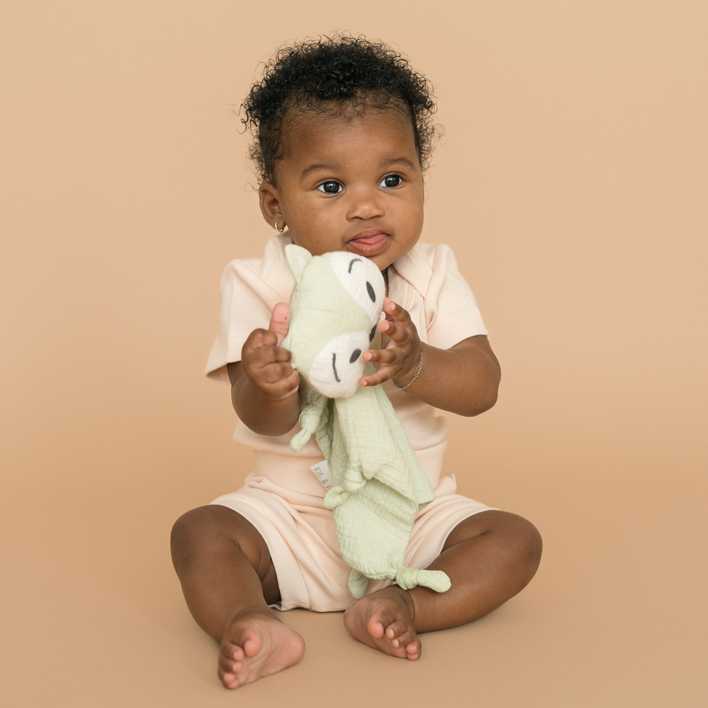 Baby girl wearing rosy peach organic cotton bodysuit and fold-over shorts, holding a green fox toy