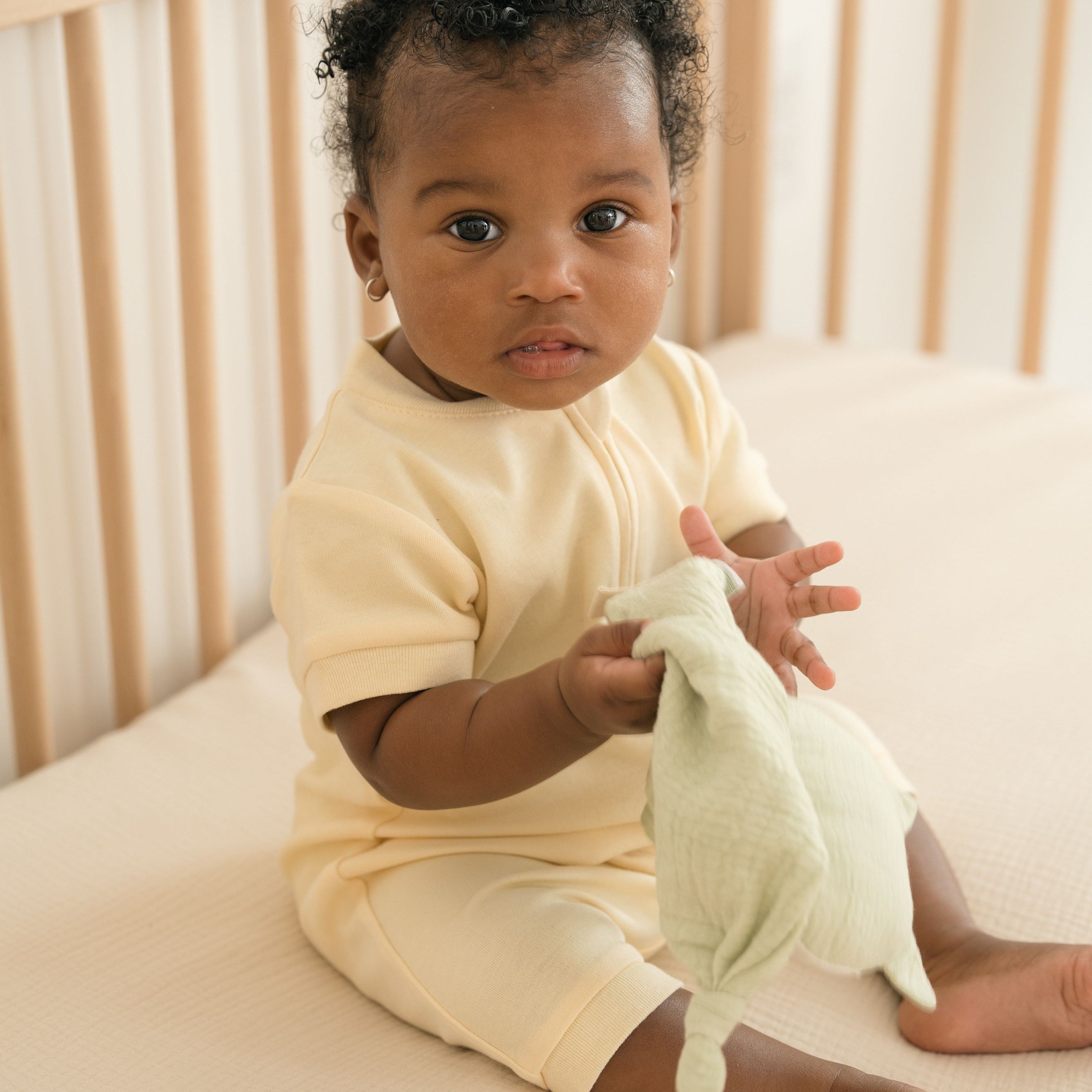 Baby sitting in a crib wearing a Sunny Yellow organic cotton short sleeve romper, holding a toy.