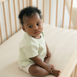 Baby sitting in a crib wearing a Leafy Green organic cotton short sleeve shirt and Forest Friends print fold-over shorts.