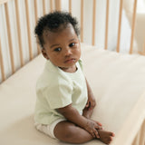 Baby sitting in a crib wearing a Leafy Green organic cotton short sleeve shirt and Forest Friends print fold-over shorts.