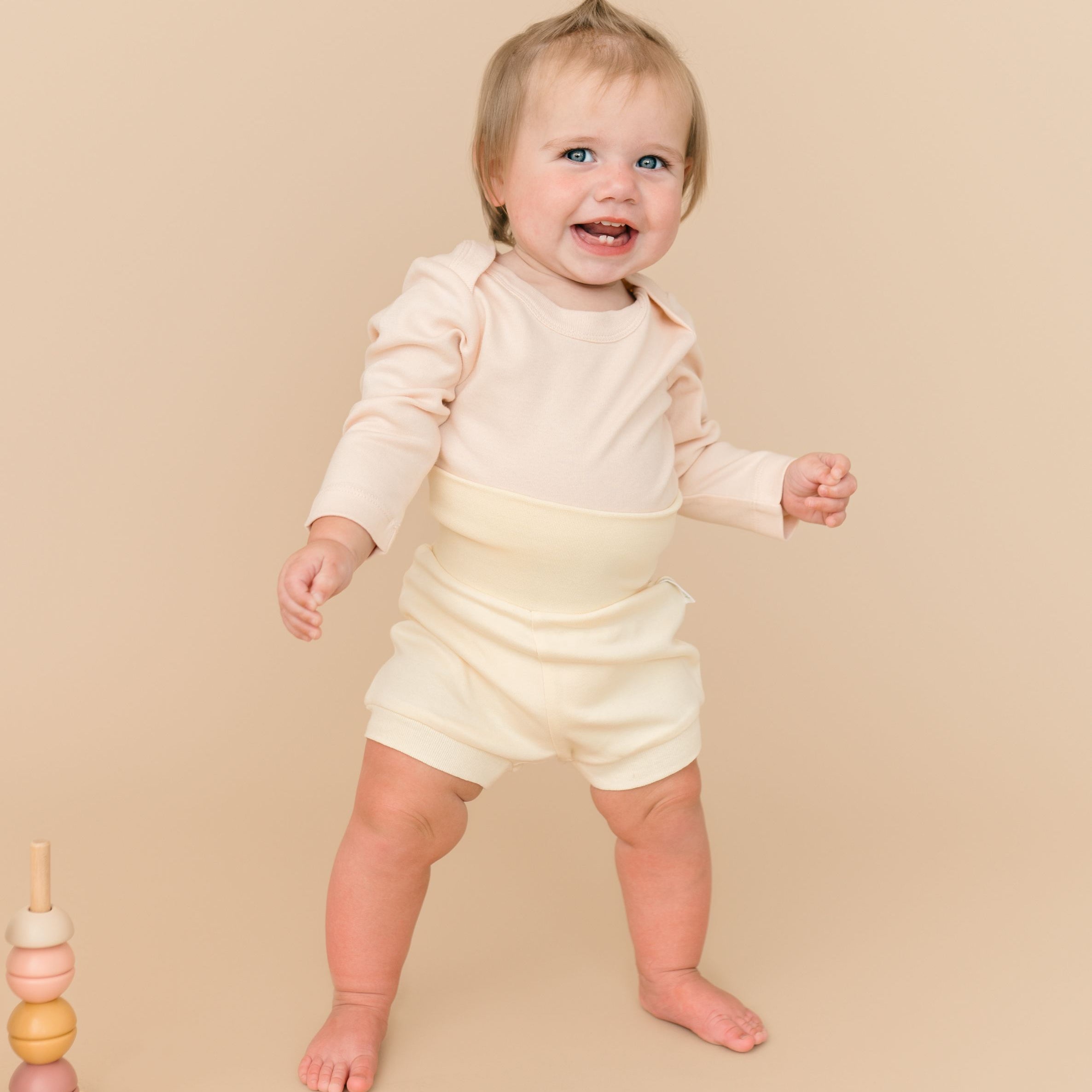 Smiling toddler girl wearing sunny yellow organic cotton fold-over shorts and rosy peach long sleeve bodysuit