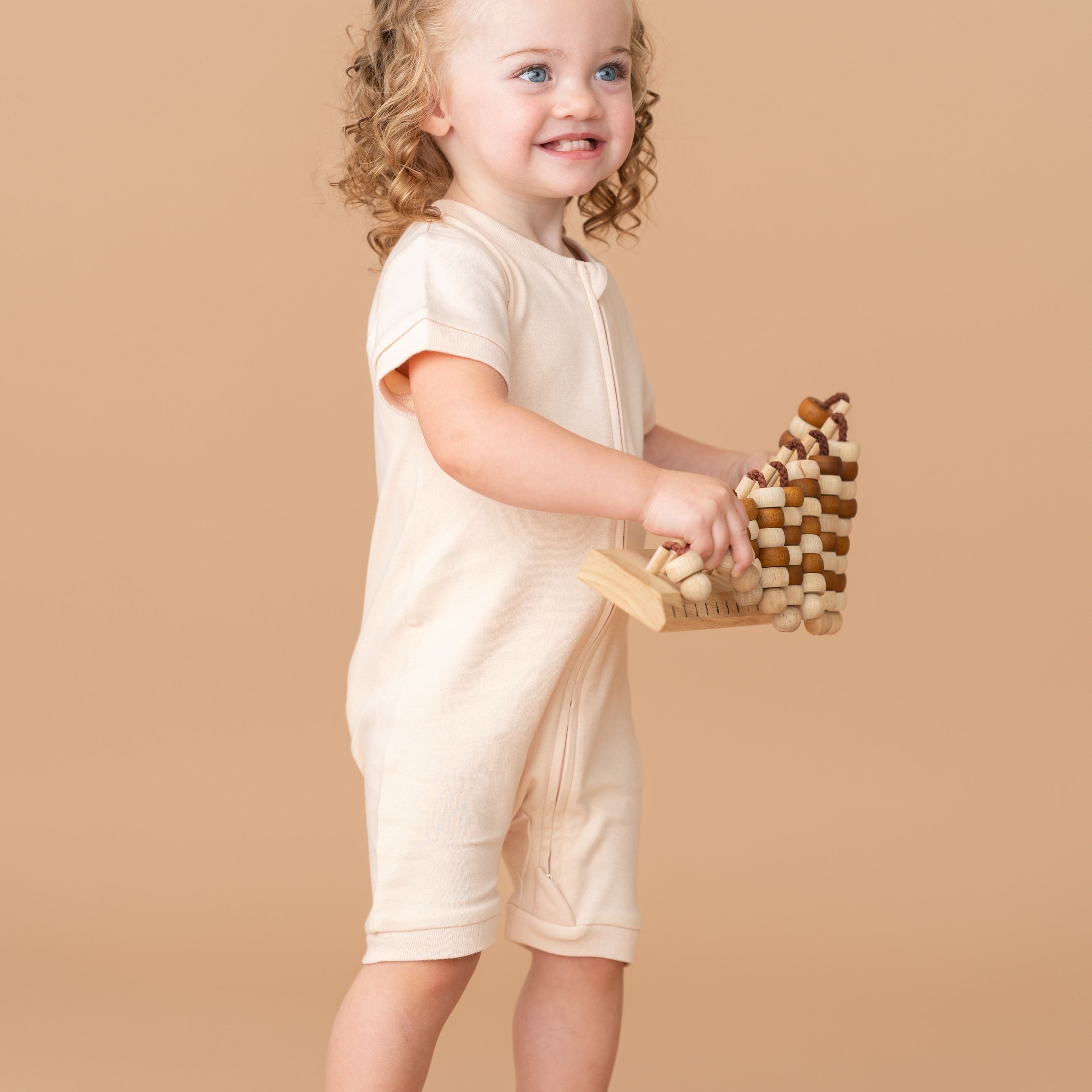 Toddler girl wearing a Rosy Peach organic cotton short sleeve romper, smiling and holding a toy while standing.