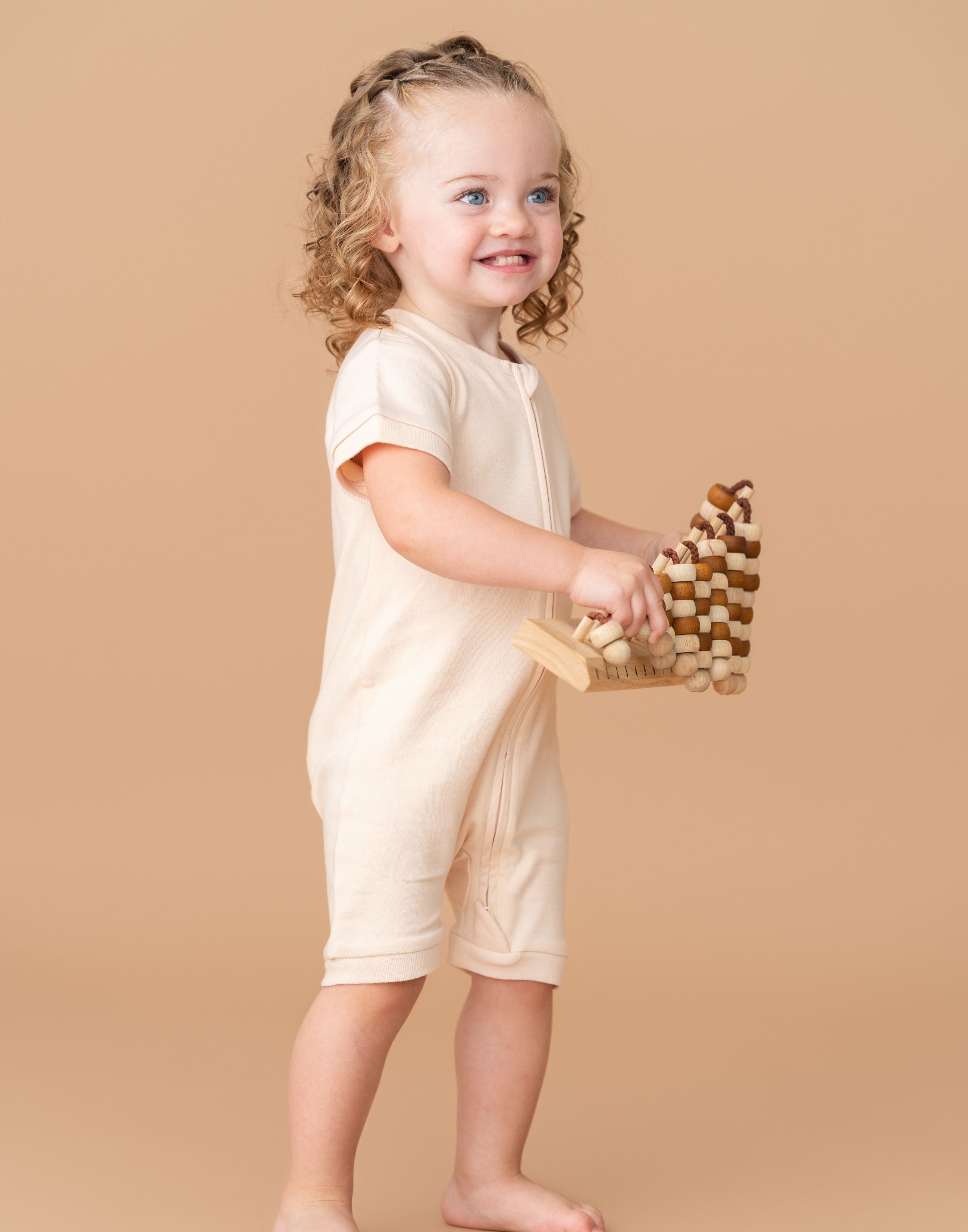 Toddler girl wearing a Rosy Peach organic cotton short sleeve romper, smiling and holding a toy while standing.