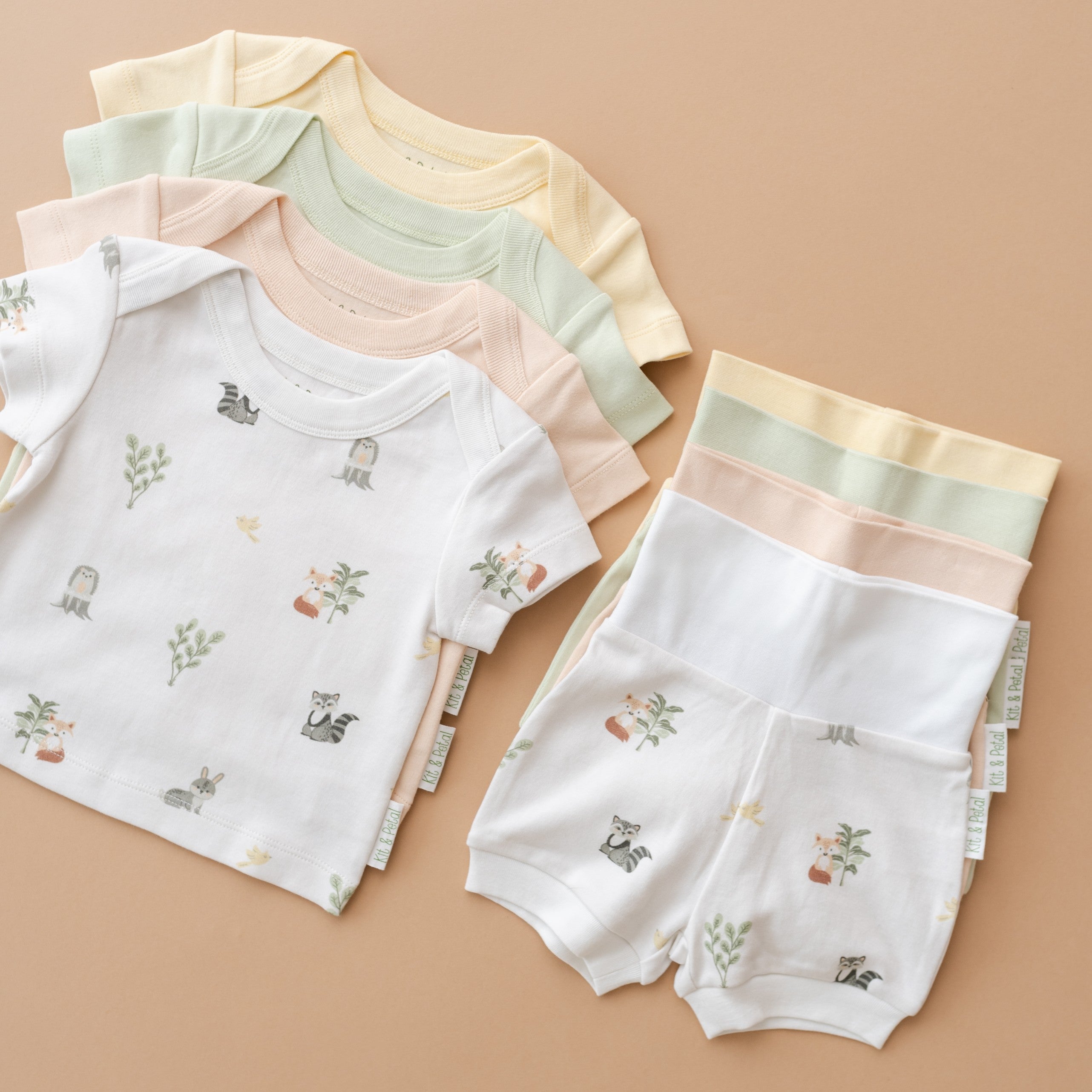 Flat lay of Kit & Petal matching organic cotton short sleeve shirts and fold-over shorts in Forest Friends, Leafy Green, Sunny Yellow, and Rosy Peach colorways.